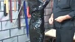 Klobige brünette BDSM-Sklavin wird von Kopf bis Fuß in schwarzen Plastik gewickelt