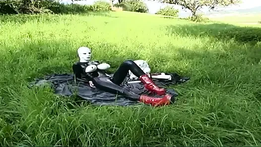 Fetish slave in latex walks and sucks a dildo on the grass