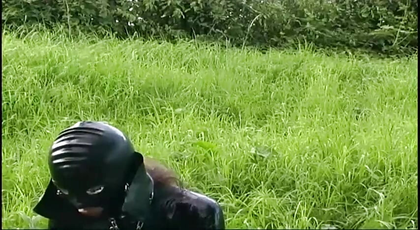 Fetish slave in latex walks and sucks a dildo on the grass