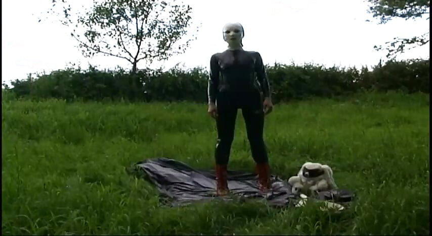 Fetish slave in latex walks and sucks a dildo on the grass
