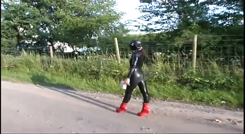 Fetish slave in latex walks and sucks a dildo on the grass