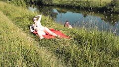 狂野的海滩。性感的熟女白金在河岸上裸体晒日光浴，随机渔夫观看。在公共场合裸体。裸体海滩