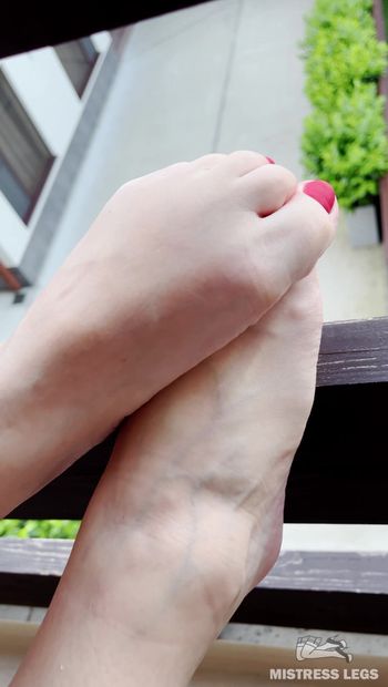 Barefoot with red toenails on the balcony. I'm sitting on the balcony and resting with my legs stretched out on the balcony railing. Matte red toenails look so luscious in the sunlight. Enjoy my toes.