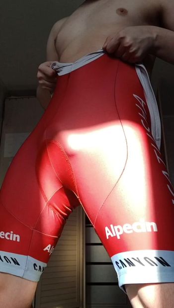 A sexy twink stands against the rays of the morning sun, his red tight cycling suit gently accentuating his slender curves. The guy self-confidently demonstrates his arousal