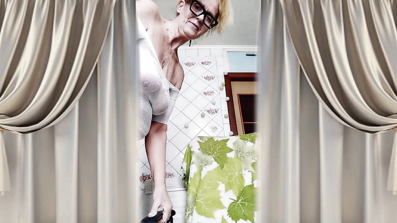 Cheerful housewife Lukerya flirts in an open, transparent white jumpsuit in the kitchen.