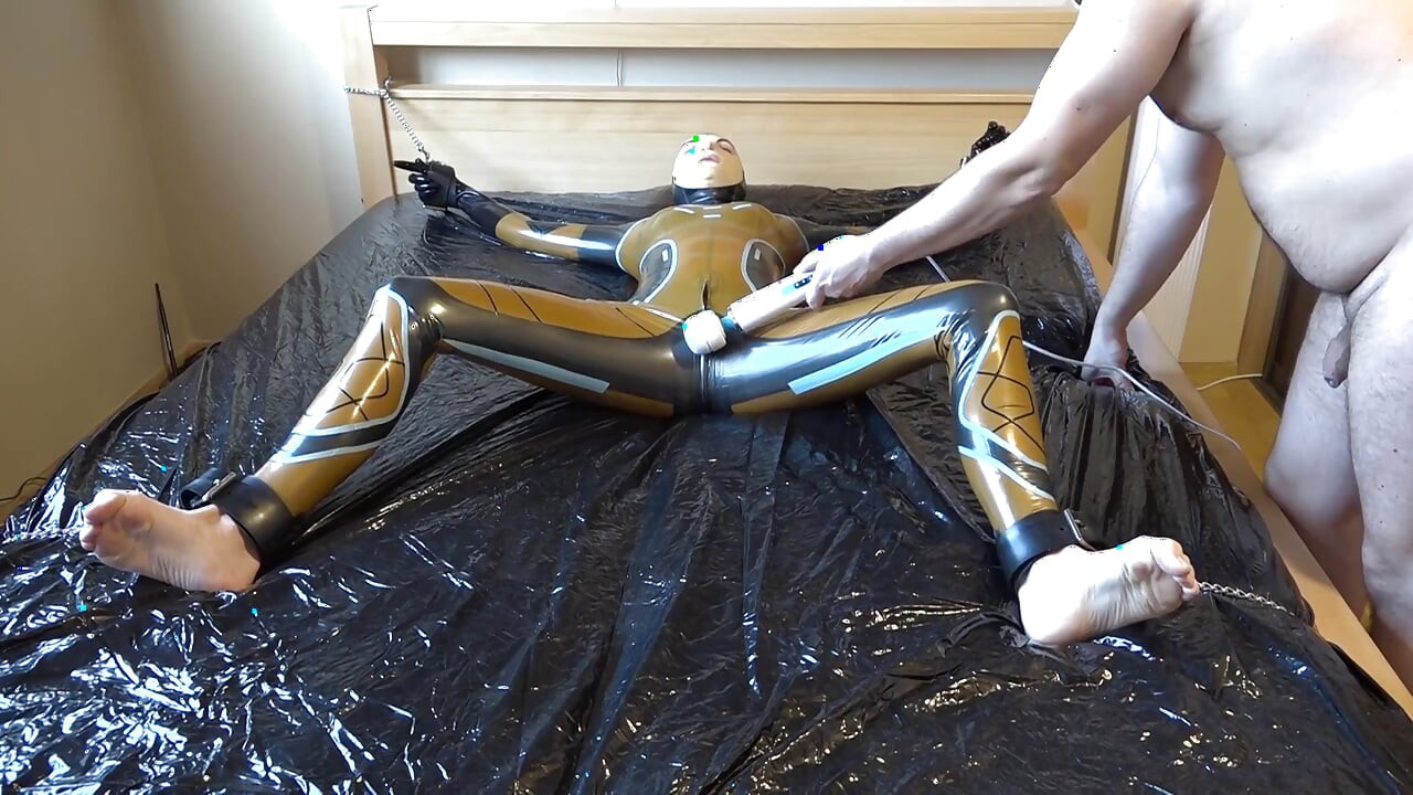 Danielle, în latex, este atașată de pat și se masturbează cu vibratorul de masaj