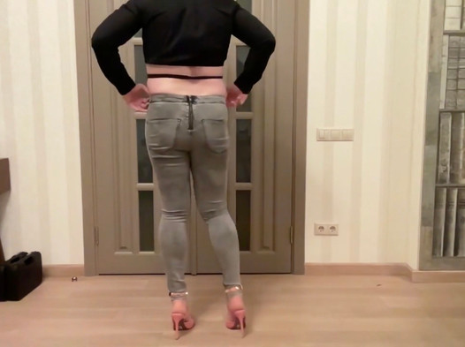 Femboy in een grijze skinny jeans met ritssluiting, hoge hakken en een zwarte bijgesneden blouse danst, masturbeert en komt klaar