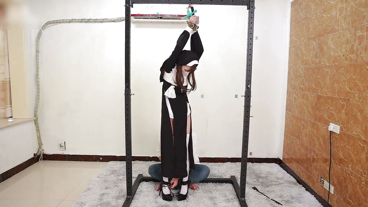 A namorada interpreta uma freira, algemada e algemada, amarrada a uma estrutura de ferro