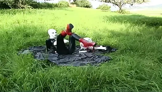Fetish slave in latex walks and sucks a dildo on the grass