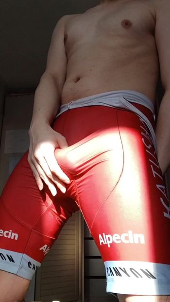 セクシーなイケメンが朝日を浴びて立ち、赤いタイトなサイクリングスーツがほっそりとした曲線を優しく強調しています。男は自信満々に興奮を示します