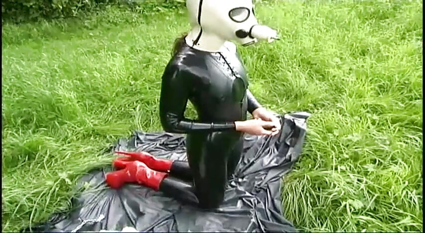 Fetish slave in latex walks and sucks a dildo on the grass