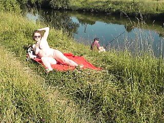 Spiaggia selvaggia. sexy milf platino nuda che prende il sole sulla riva del fiume, guarda un pescatore casuale. nudo in pubblico. spiaggia nudista