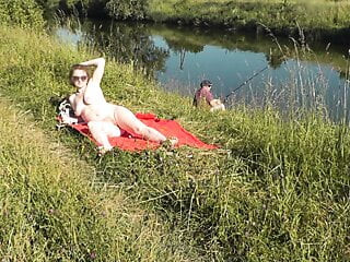 Plage sauvage. Une MILF sexy platine se fait bronzer nue sur la berge de la rivière, un pêcheur regarde au hasard. nue en public. plage naturiste