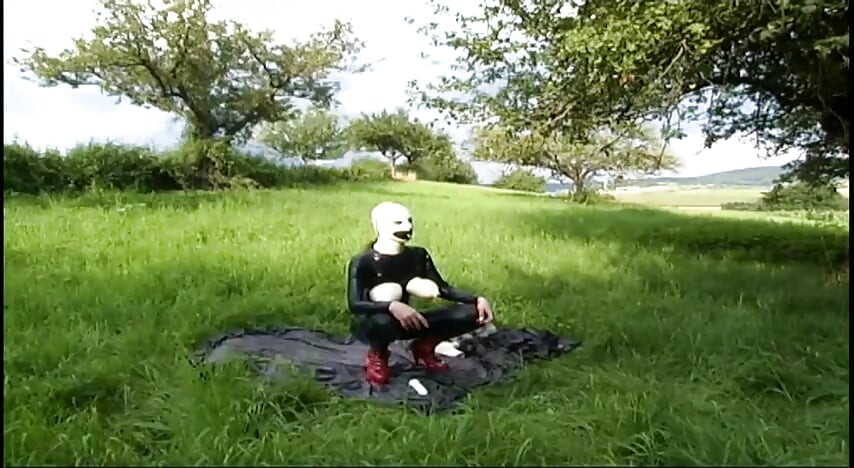 Fetish slave in latex walks and sucks a dildo on the grass