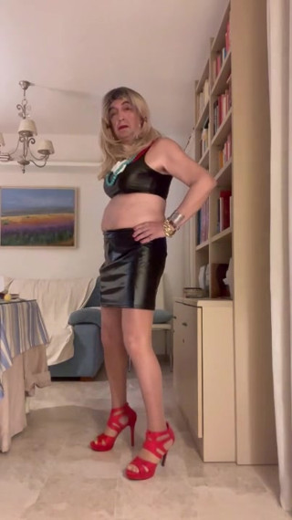 Beautiful Spanish trans woman poses seductively in the living room wearing a black latex miniskirt and red high heels thumbnail